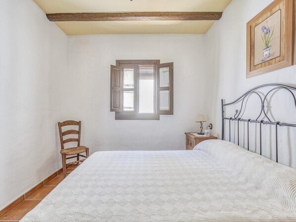 Un dormitorio con cama, una silla de madera, una mesita de noche con lámpara y una ventana con contraventanas abiertas.