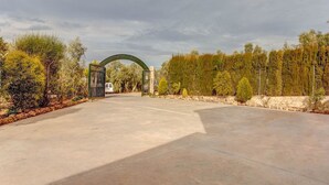 Une allée pavée mène à une entrée fermée par un portail, avec un mur de pierres et des arbres de chaque côté.