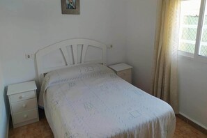 Un dormitorio con cama, dos mesitas de noche, una mesilla y una ventana con cortinas, además de un cuadro colgado en la pared.