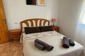 A neatly made bed with a patterned bedspread and two rolled towels, a wooden nightstand with a lamp, and a door to the left.