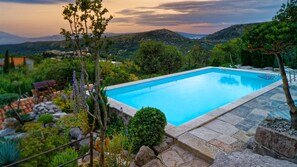 Una piscina rectangular con borde de piedra, rodeada de vegetación y un paisaje montañoso.