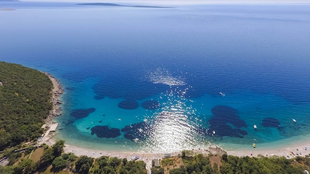 Vista aérea de una zona costera con aguas azules cristalinas, formaciones rocosas y una playa con gente y barcos.