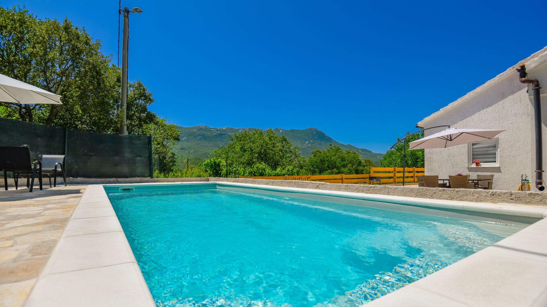 A swimming pool with clear blue water, surrounded by a patio area with outdoor furniture, a mountain view, and a white building with a window.