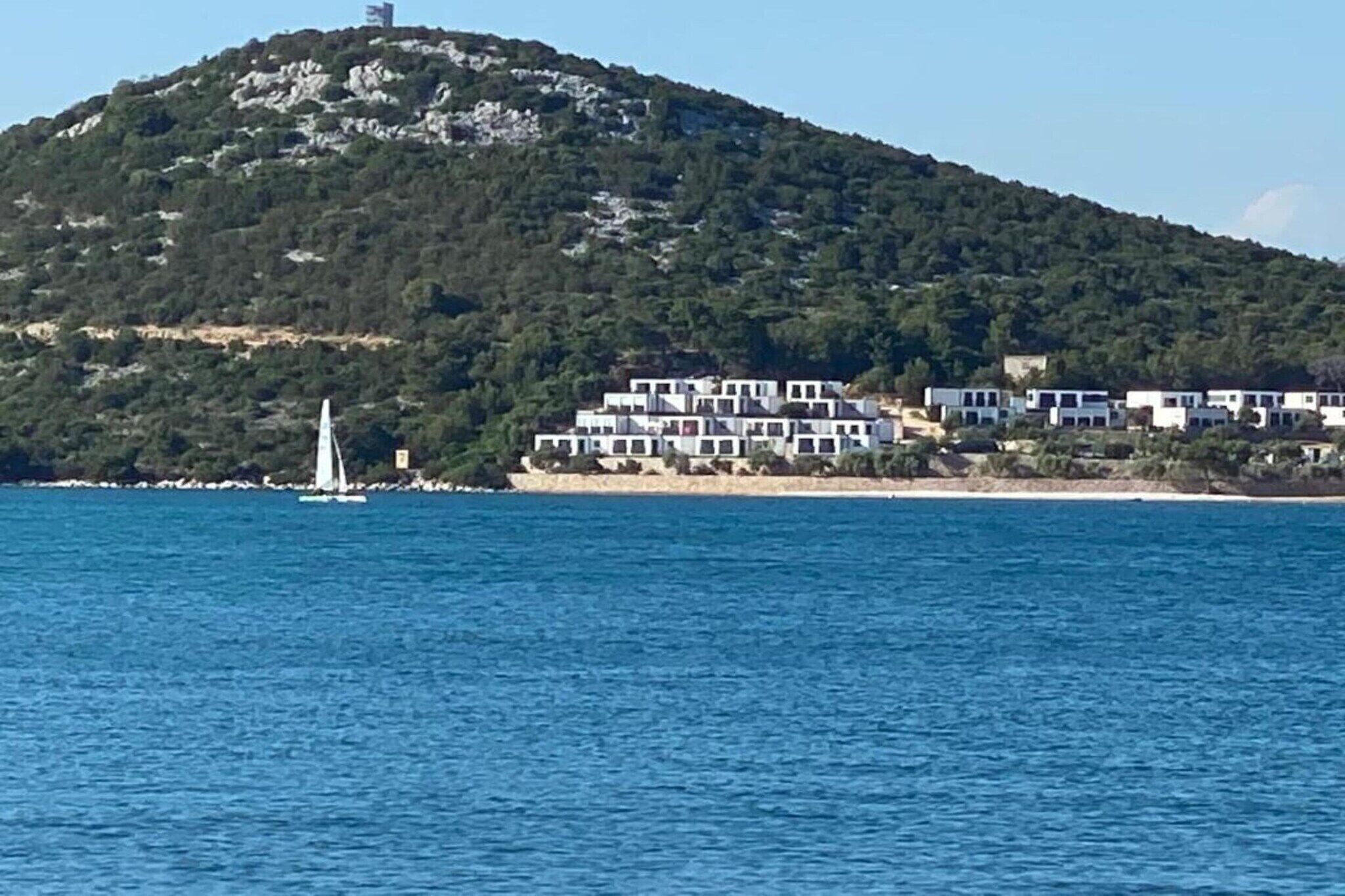 Ein Küstenkomplex mit mehreren weißen Gebäuden, einem Segelboot im Wasser und einem Hügel mit spärlicher Vegetation.