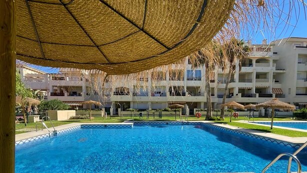 A swimming pool with a thatched umbrella, surrounded by a poolside area with a view of a multi-story building in the background.