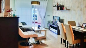 A modern living room with a sofa, coffee table, and dining area. There is a TV on a stand and a bookshelf with decorative items.