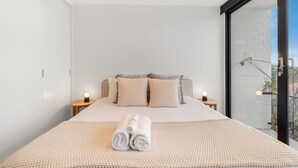 A modern bedroom with a large bed, two bedside tables, and a view of a cityscape through a large window.