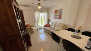 A living room with a sofa, a dining table with chairs, a ceiling fan, and a window with curtains.