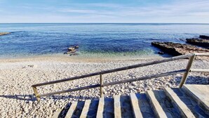 Une plage de galets avec un escalier en bois descendant jusqu’à l’eau, un ciel dégagé et une mer calme.