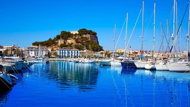 Un port de plaisance avec des bateaux amarrés, une colline parsemée de bâtiments et un ciel bleu dégagé.