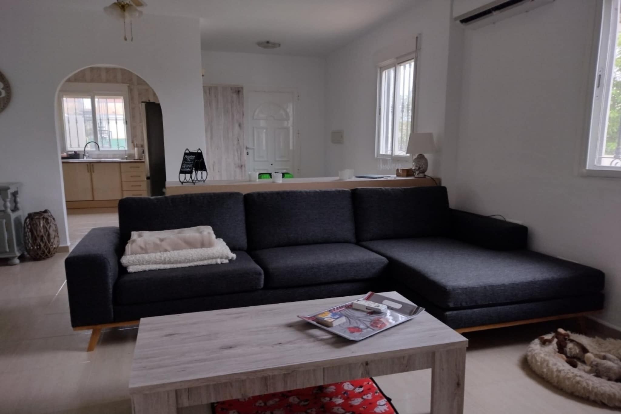 Un salon moderne avec un canapé d’angle gris foncé, une table basse en bois et un panier pour animaux de compagnie. On aperçoit une cuisine à travers une arche en arrière-plan.