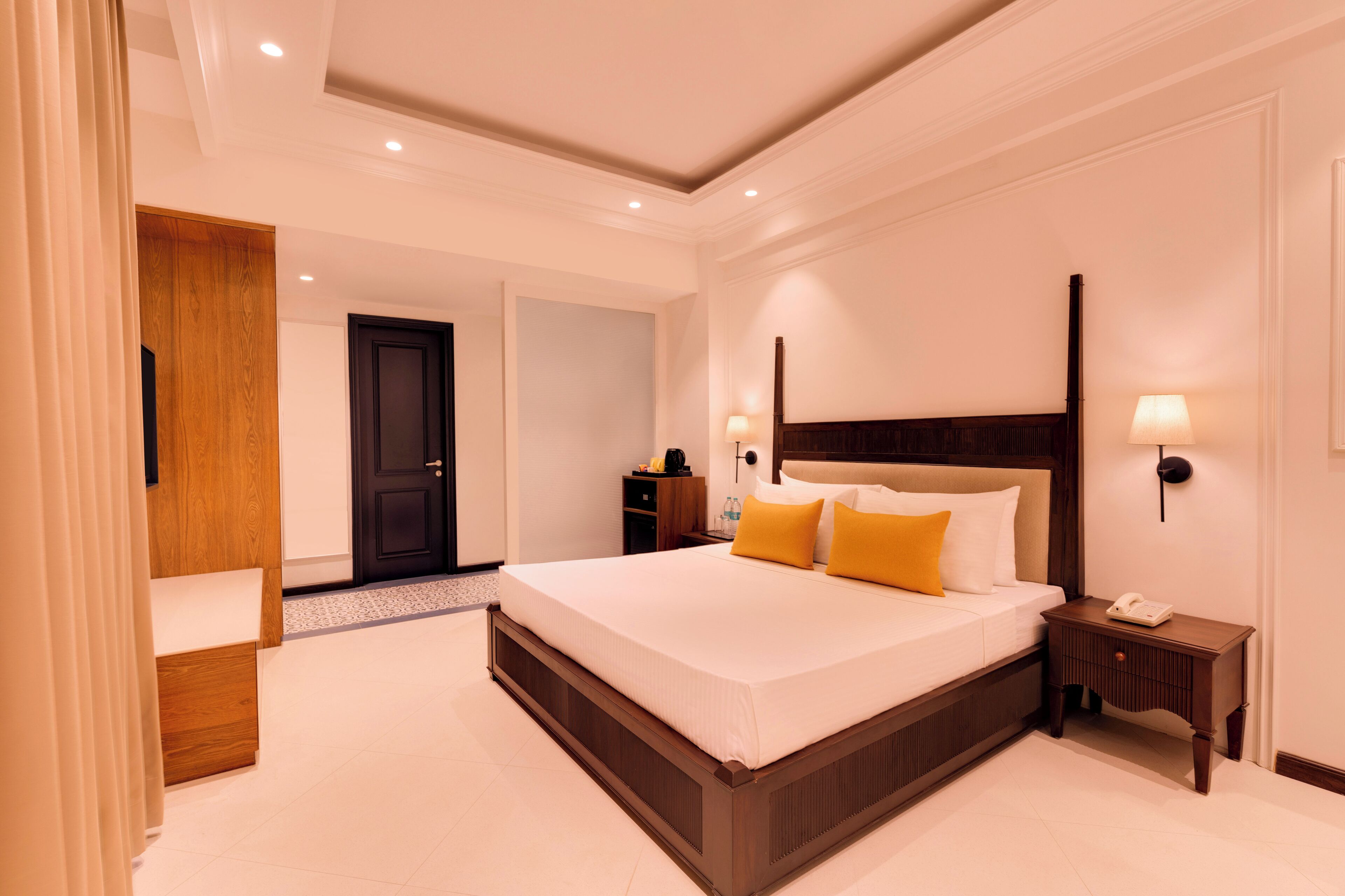 A hotel room with a large bed, bedside tables, a wooden wardrobe, and a door leading to another room.