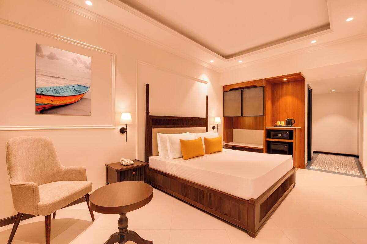 Habitación de hotel con una cama grande, una silla, una mesita, un cuadro en la pared y una zona de kitchenette.