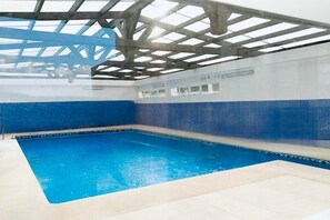 Une piscine intérieure avec un mur recouvert de carreaux bleus et un plafond doté d’une structure métallique.
