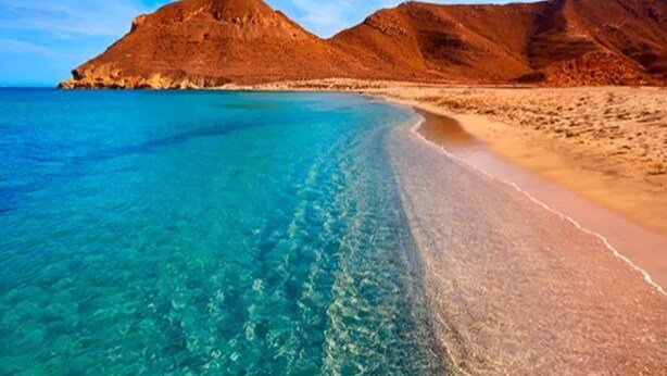 A beach with clear turquoise water and sandy shores, surrounded by reddish-brown hills.