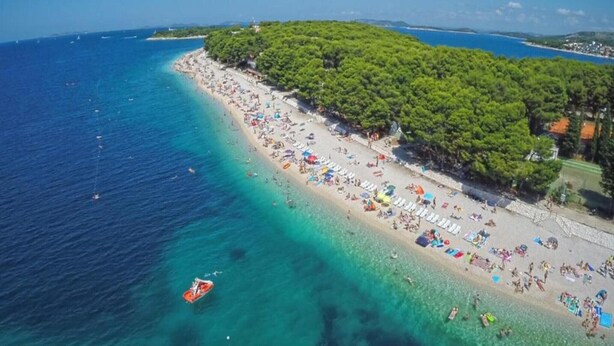 An aerial view of a beach with clear turquoise waters, a sandy shore, and dense green trees on the right. - Beautiful apartment near Rtić beach (Primošten)