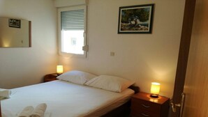 A neatly made bed with white linens and a rolled towel on the side, a bedside lamp, a small wooden nightstand, a framed picture on the wall, and a window with blinds.