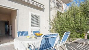Een terras met een tafel, stoelen, een raam met luiken en een balkon met een hekwerk.