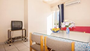 Een eetgedeelte met een glazen tafel, stoelen, een televisie op een standaard, een schaal met fruit en een vaas met bloemen.
