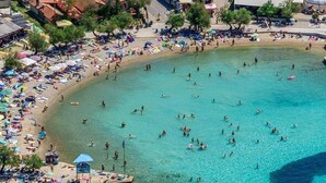 Een druk strand vol mensen die zwemmen en zonnebaden, omgeven door gebouwen en bomen.