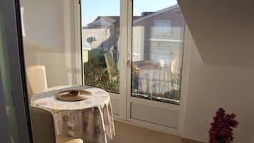 A small balcony with a table and chair, a potted plant, and a view of buildings and greenery.