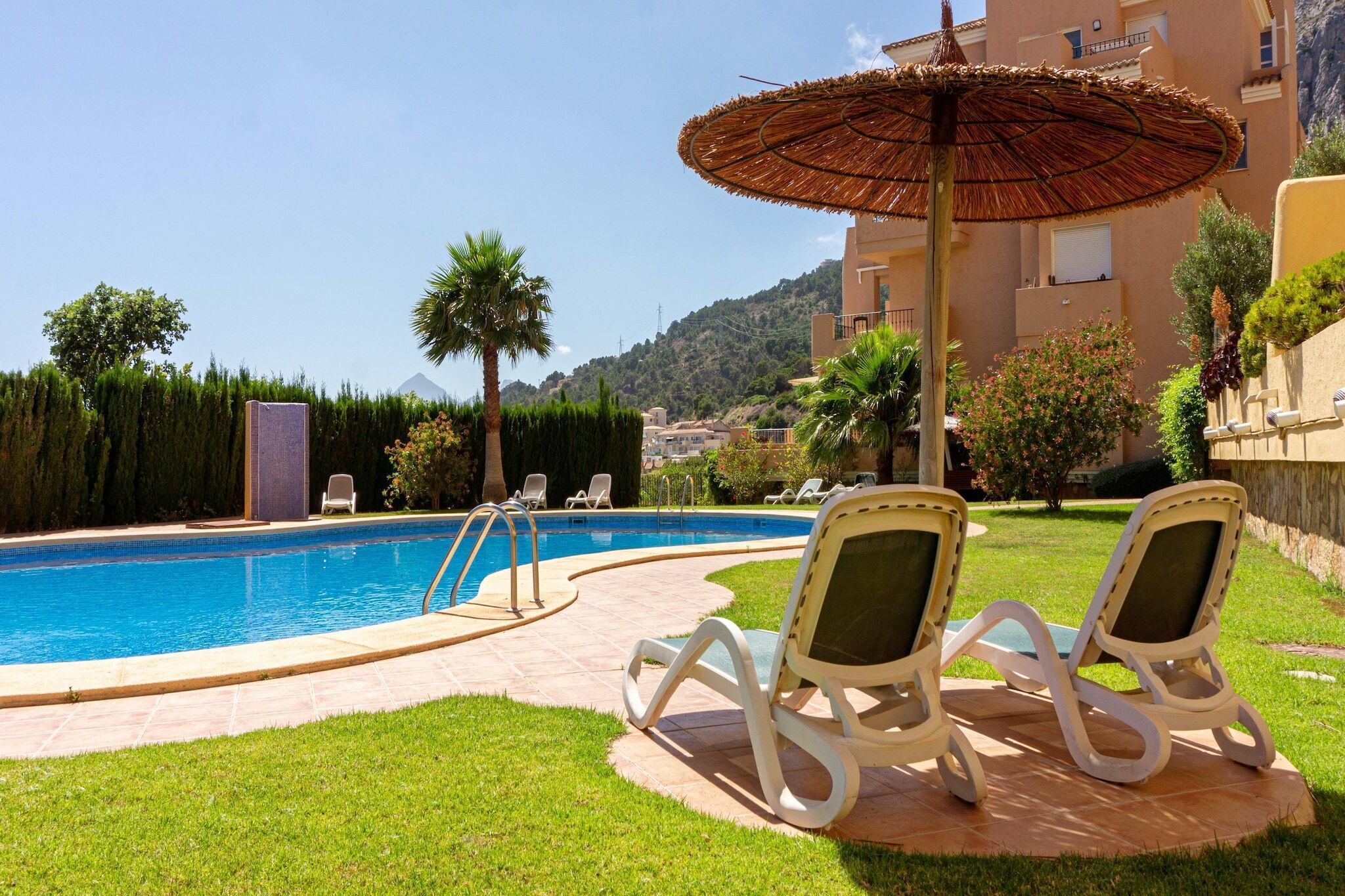 A pool area with lounge chairs and an umbrella, surrounded by greenery and a building in the background.