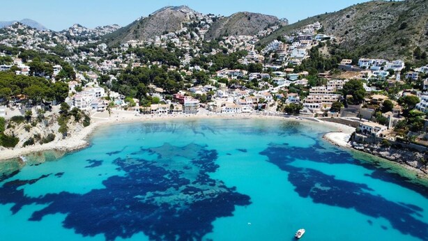 A coastal town with a beach, clear blue waters, and buildings on a hillside.