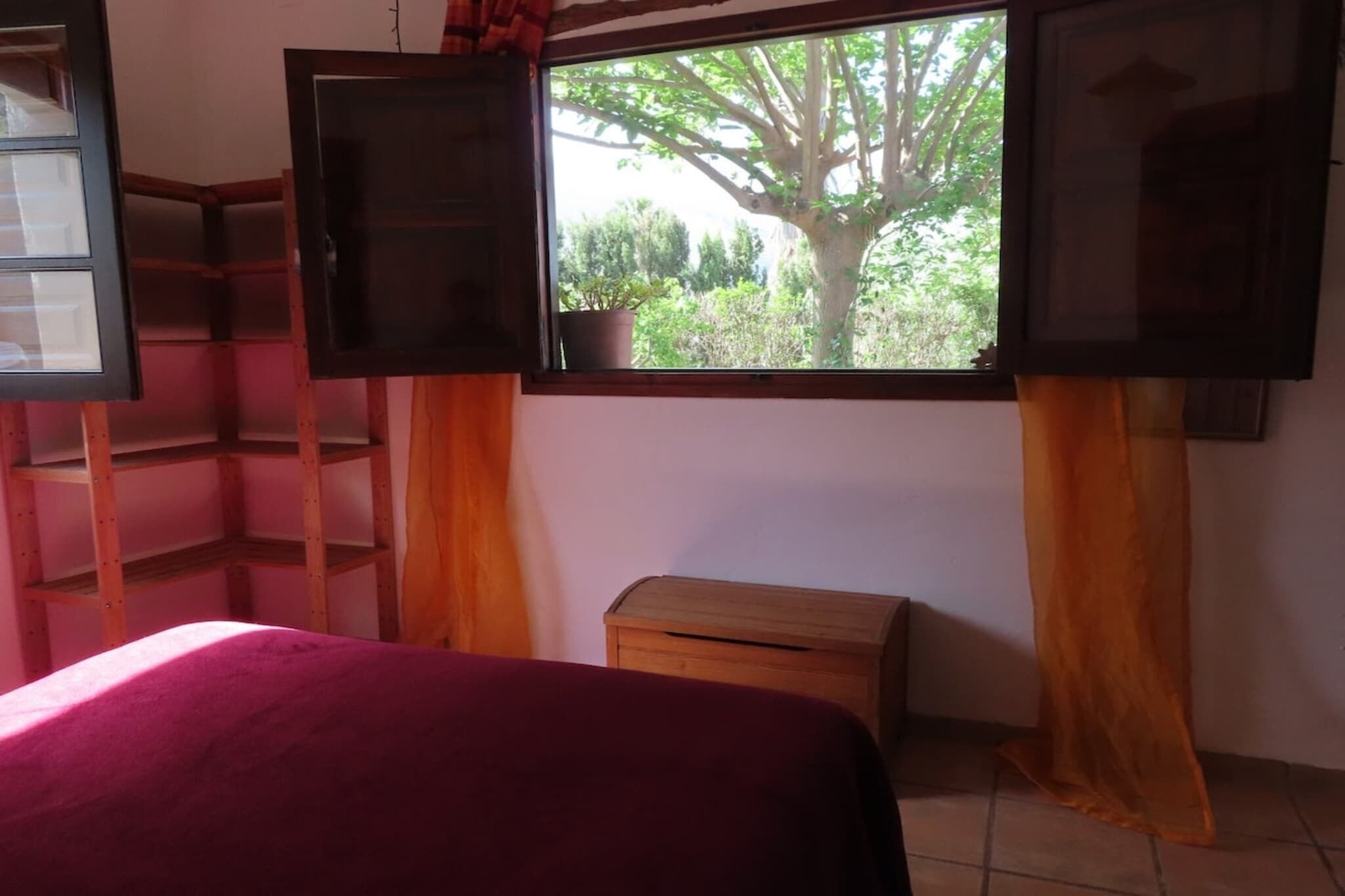 A room with a bed, a wooden shelf, a small wooden chest, and a window with a view of trees and a potted plant.