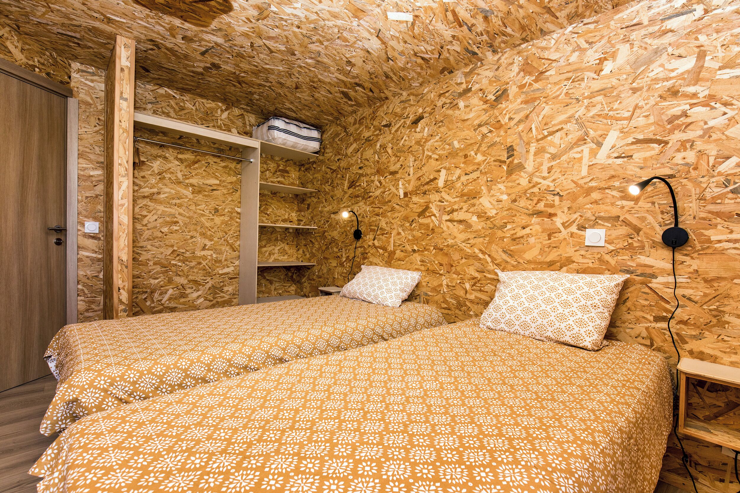 Una habitación con dos camas, paredes de madera, un armario, una estantería con una bolsa y una lámpara de pared.