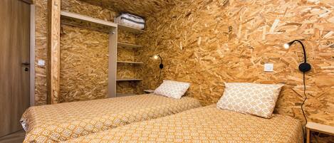 Una habitación con dos camas, paredes de madera, un armario, una estantería con una bolsa y una lámpara de pared.