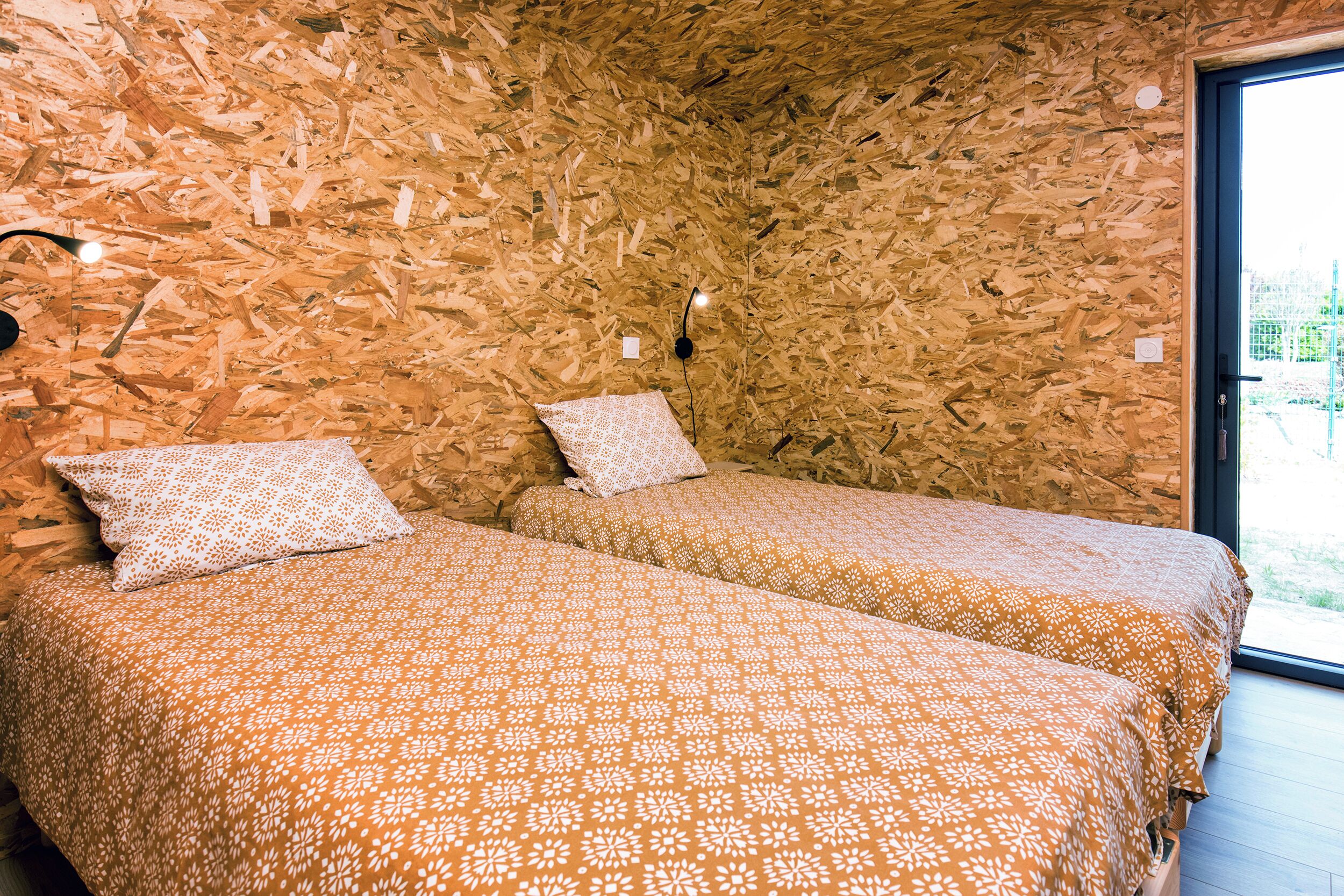 A room with two beds, each with patterned bedding, a wooden wall, and a window.