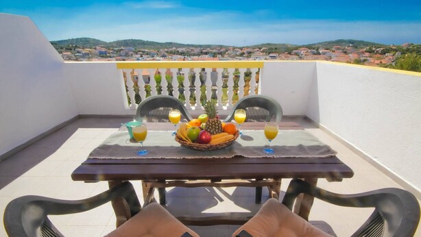 Een balkon met een tafel gedekt voor twee personen, met een fruitschaal en twee glazen sinaasappelsap.