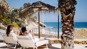 A beachside lounge with white lounge chairs, a palm tree, and a view of the sea and buildings in the background.