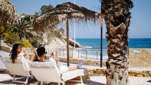 A beachside lounge with white lounge chairs, a palm tree, and a view of the sea and buildings in the background.