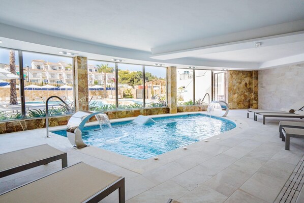 Una piscina interior moderna con cascada, amplias ventanas que ofrecen vistas de los edificios y la vegetación circundantes.