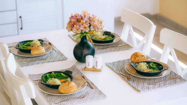 Une table à manger dressée avec des assiettes remplies de nourriture, un vase de fleurs, et des salières et poivrières.