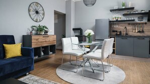 Un salon moderne avec un canapé bleu, une table à manger avec des chaises blanches, un coin cuisine avec des meubles gris et une grande horloge murale.