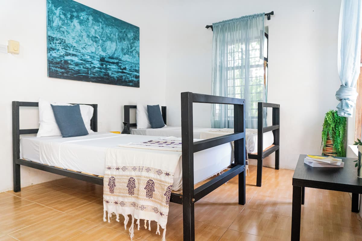 A bedroom with a bed, a bedside table, a window with curtains, a painting on the wall, and a potted plant.