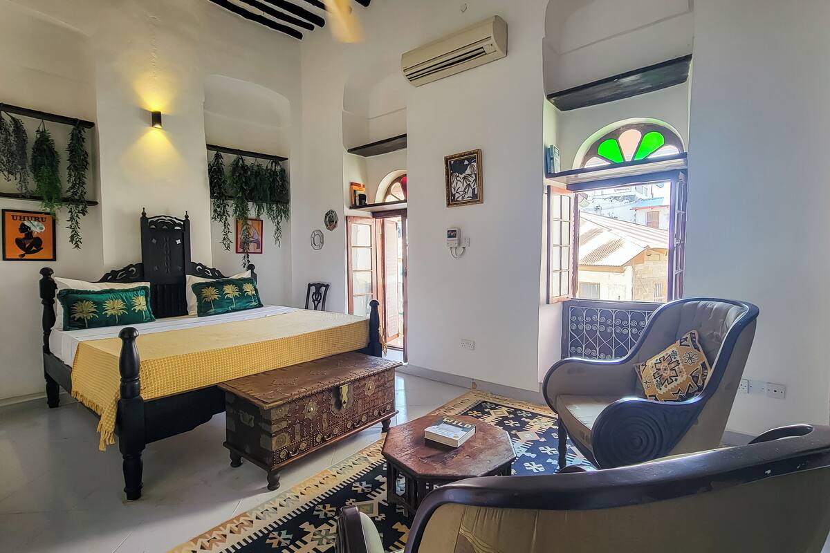 A room with a bed, a chair, a small table, and a window with a view of buildings outside.