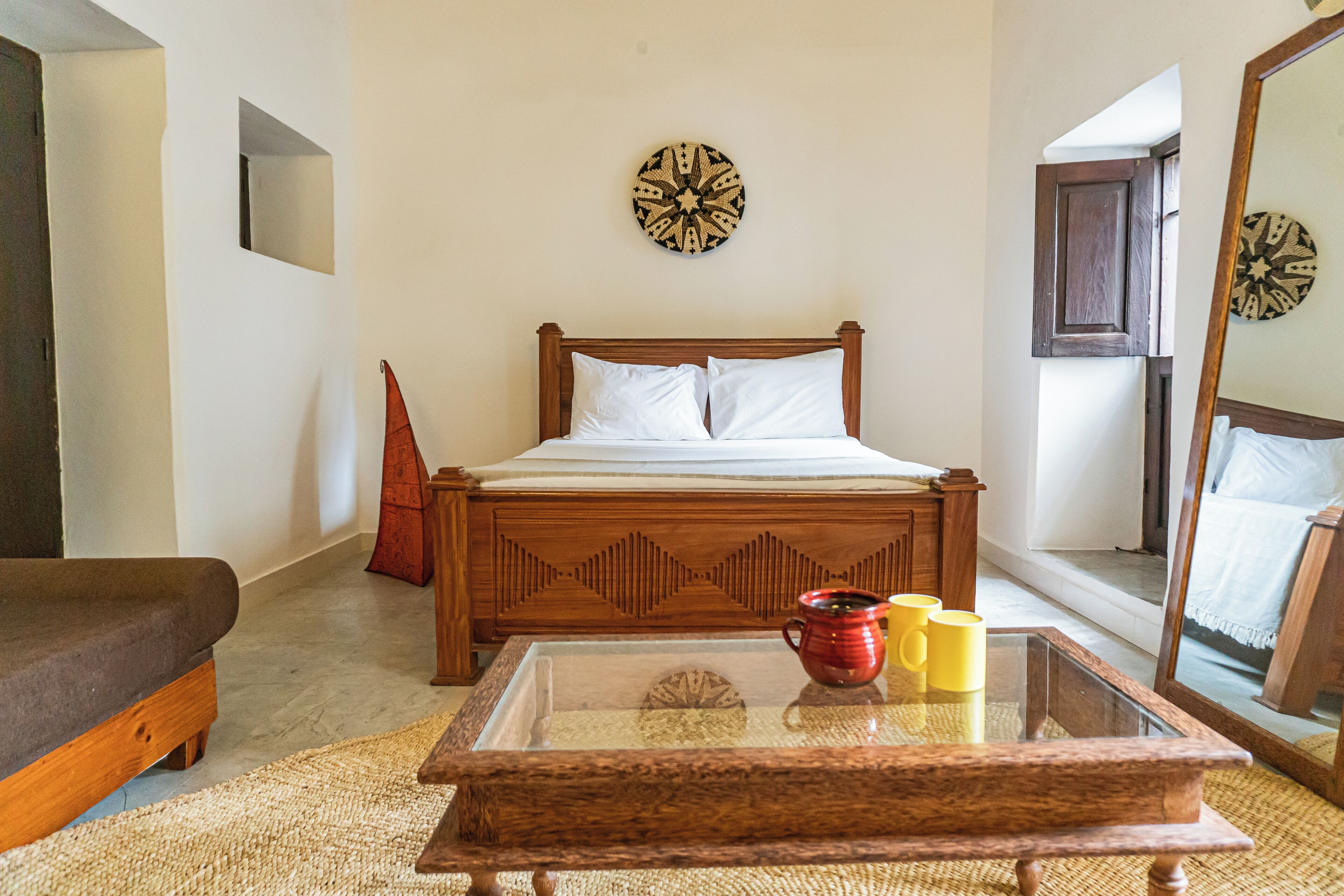 A bedroom with a wooden bed, a glass coffee table with mugs, and a mirror reflecting the room.