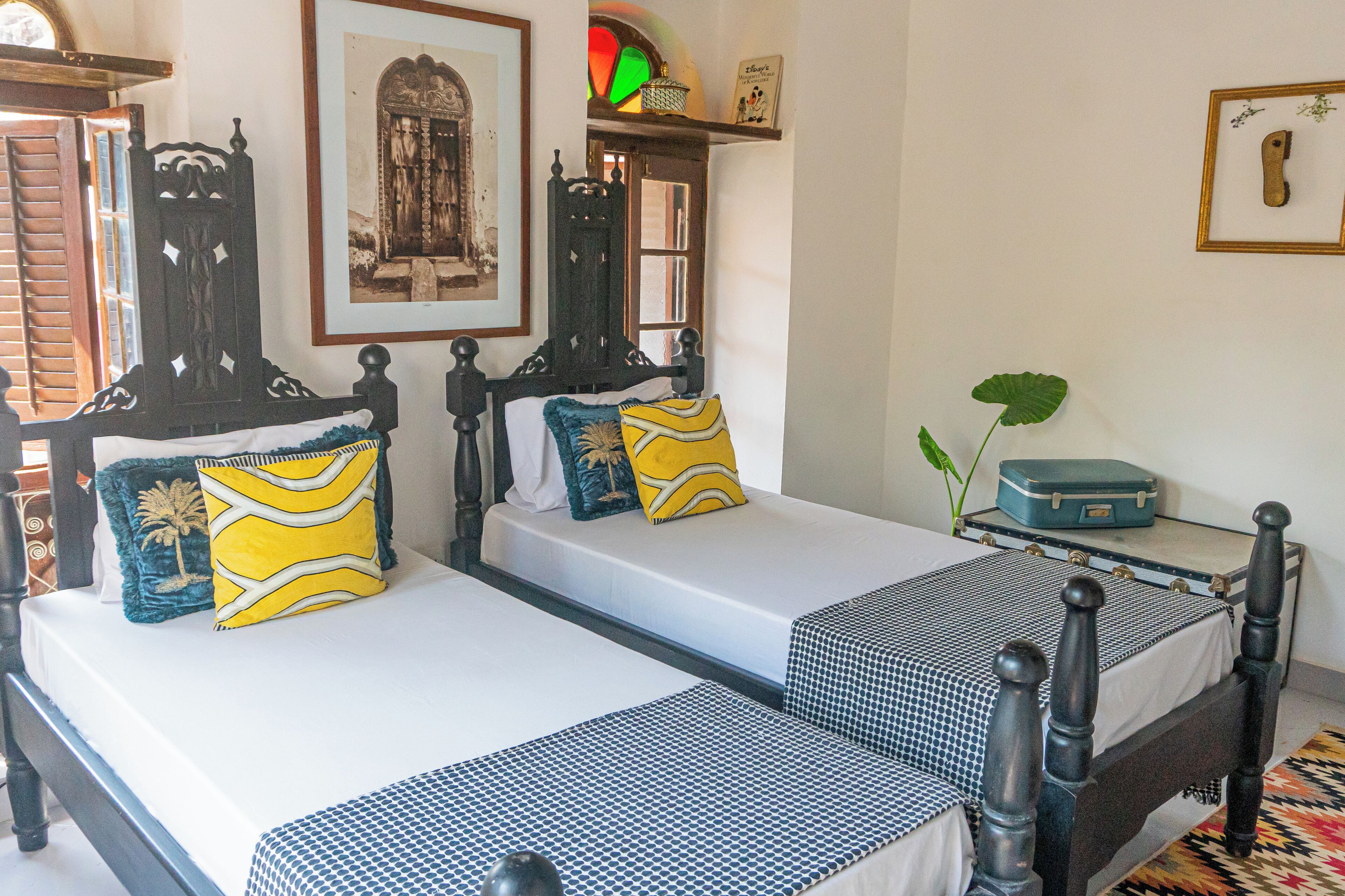 A room with two beds, each with a different patterned bedspread. There are decorative pillows, a small table with a suitcase, and a framed picture on the wall.