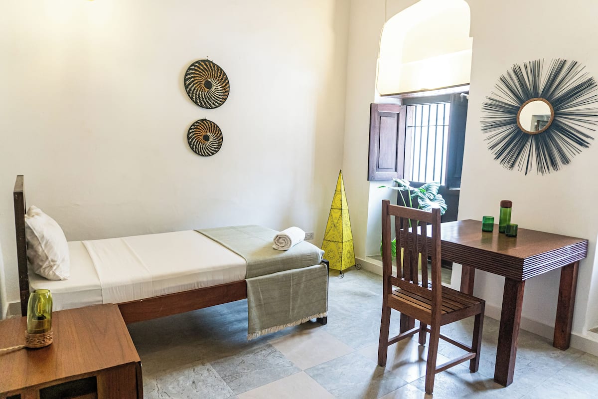 A small, well-lit room with a bed, a wooden table, and a chair. There are decorative wall art pieces and a window with bars.