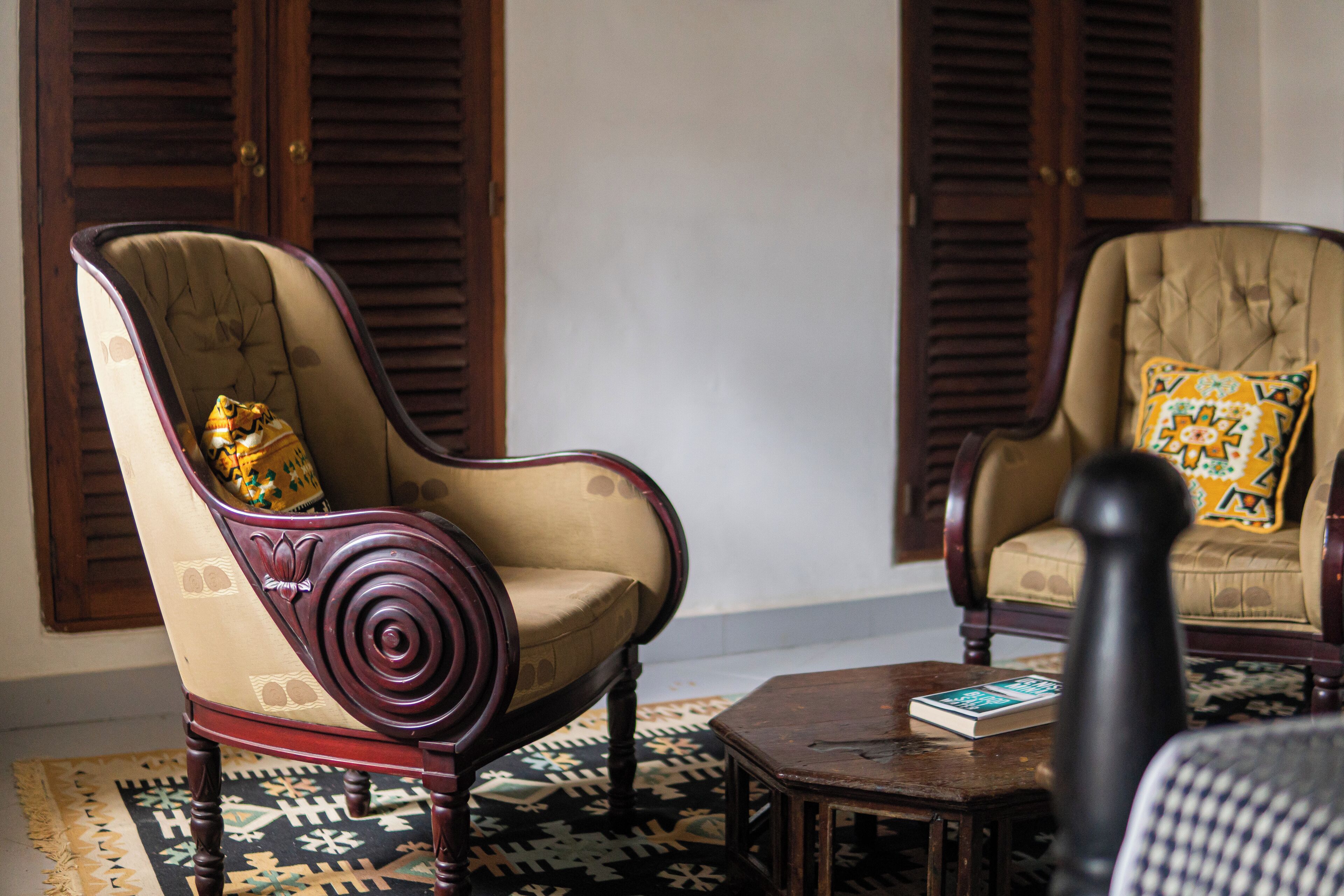Dos sillones tapizados con armazones de madera y cojines estampados, una mesita de madera con un libro, y una alfombra estampada sobre suelo de baldosas.