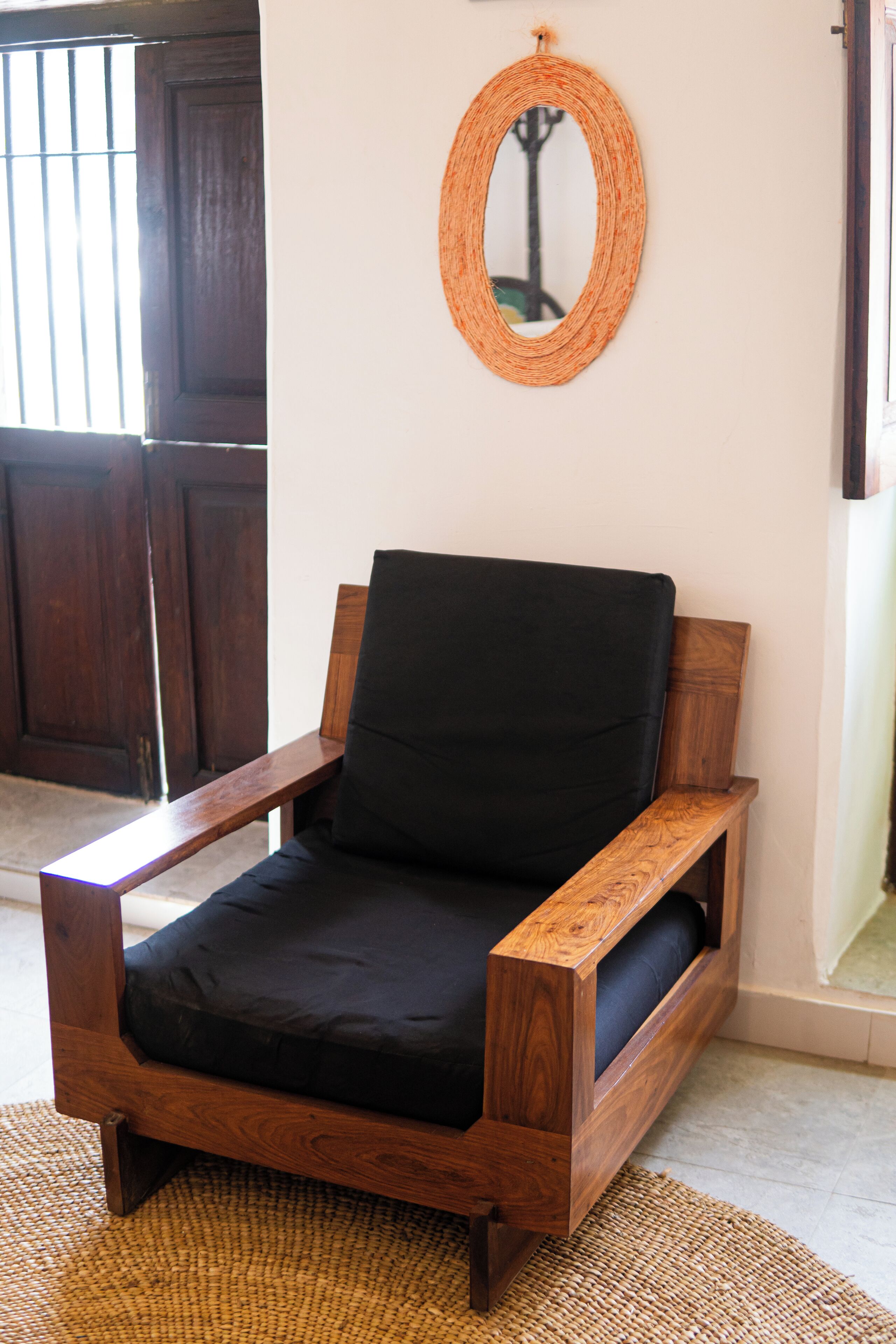 Un fauteuil en bois avec un coussin noir, un miroir tressé accroché au mur et une porte en bois à l’arrière-plan.
