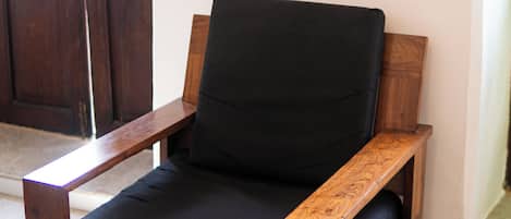 Un fauteuil en bois avec un coussin noir, un miroir tressé accroché au mur et une porte en bois à l’arrière-plan.