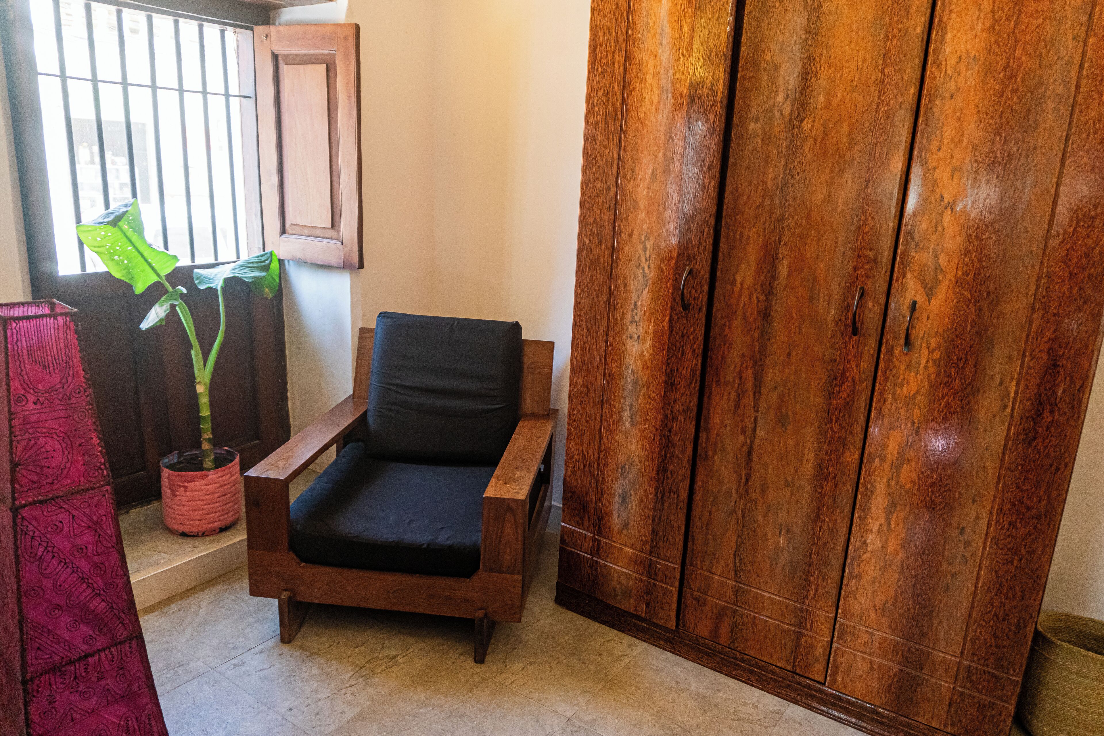 Une pièce avec une chaise en bois, une plante en pot, une armoire et une fenêtre avec des barreaux.