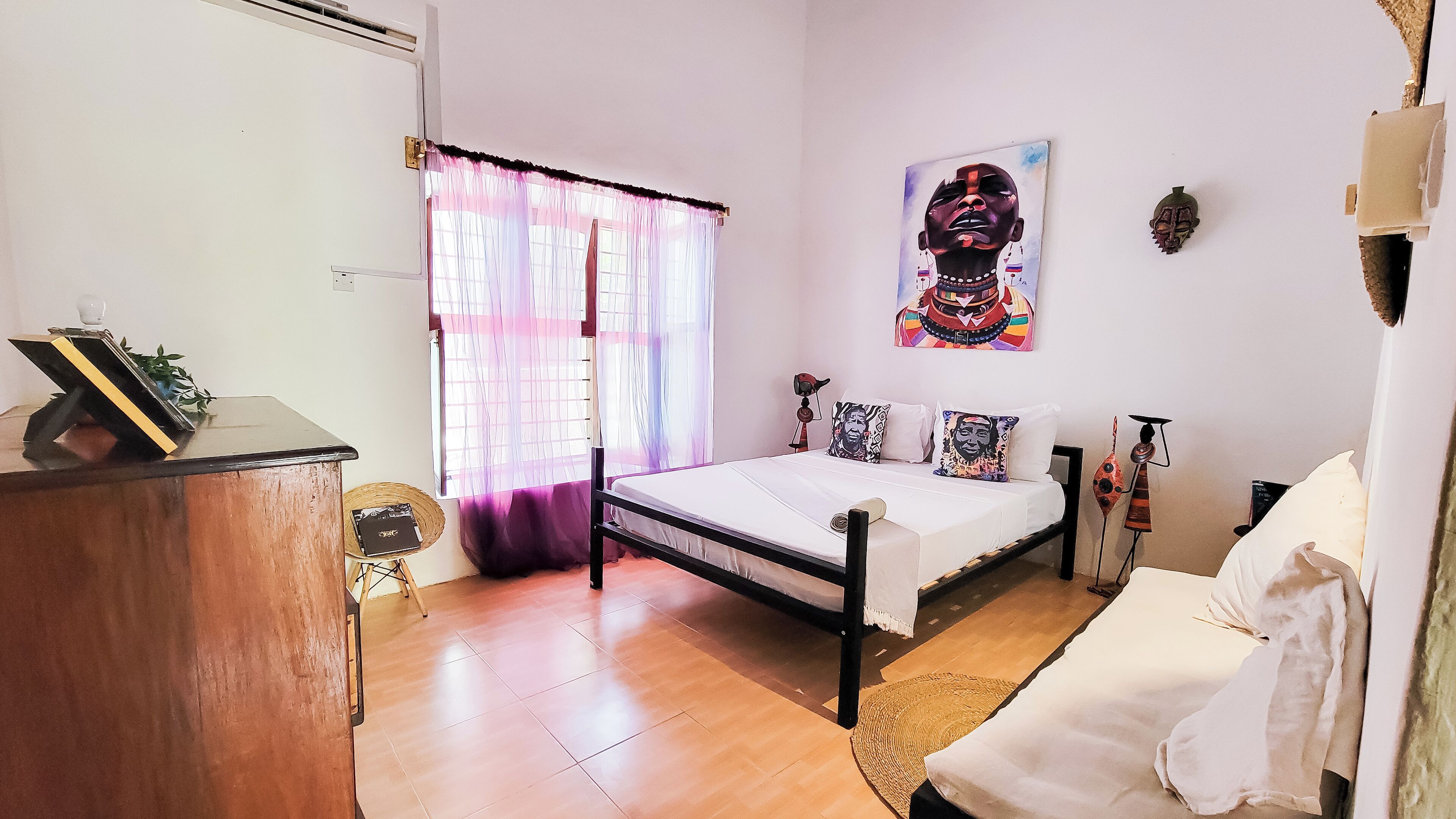 A bedroom with a bed, a bedside table, a chair, a window with curtains, a wall-mounted artwork, and a small plant.
