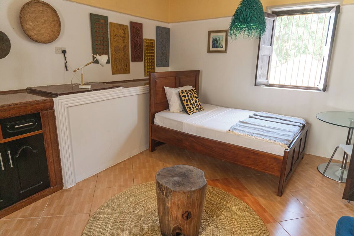 A bedroom with a wooden bed, a small bedside table, a lamp, and a window with bars.