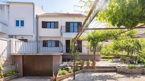 A residential building with a gated entrance, a balcony with a metal railing, and a garden area with potted plants and a bench. - 2 persons apartment with garden (Šibenik-Brodarica)