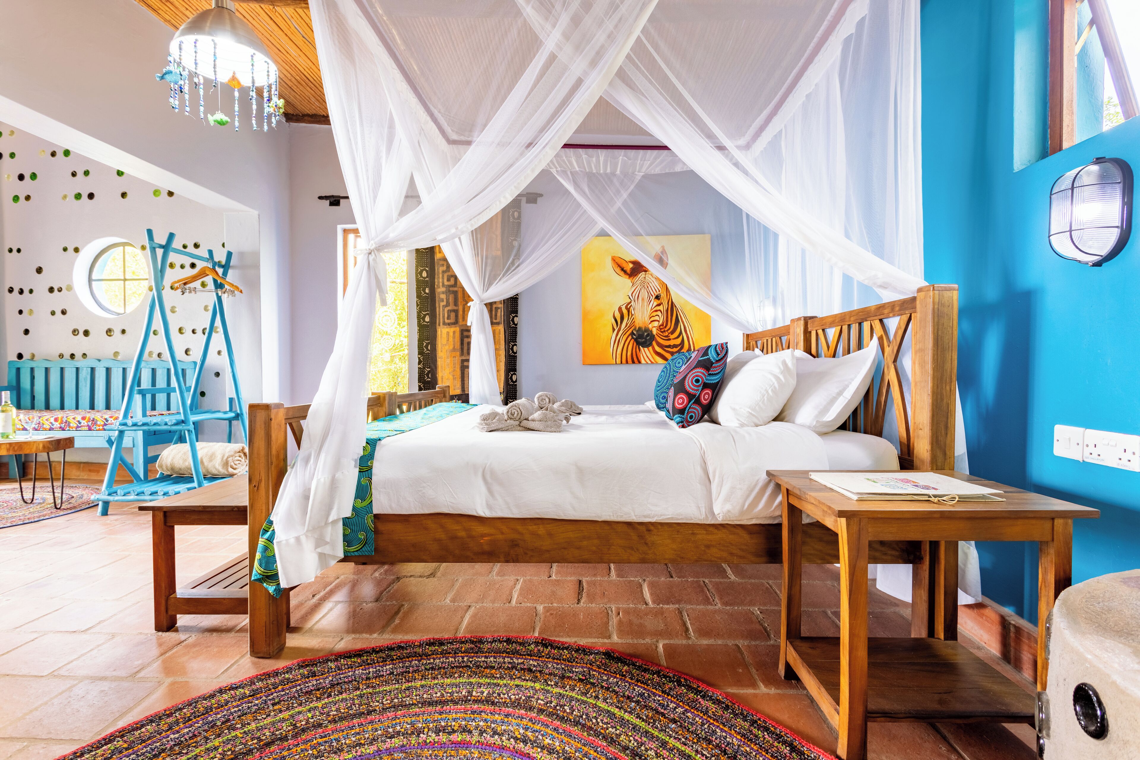 Un dormitorio con cama, una mesita de noche de madera, un tapete tejido de colores, una silla azul y un cuadro de cebra en la pared.